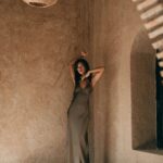 An elegant woman in a minimalist, earthy-toned interior, wearing a flowing olive-green dress. She strikes an expressive pose, surrounded by textured plaster walls and natural materials, evoking a serene and timeless aesthetic.