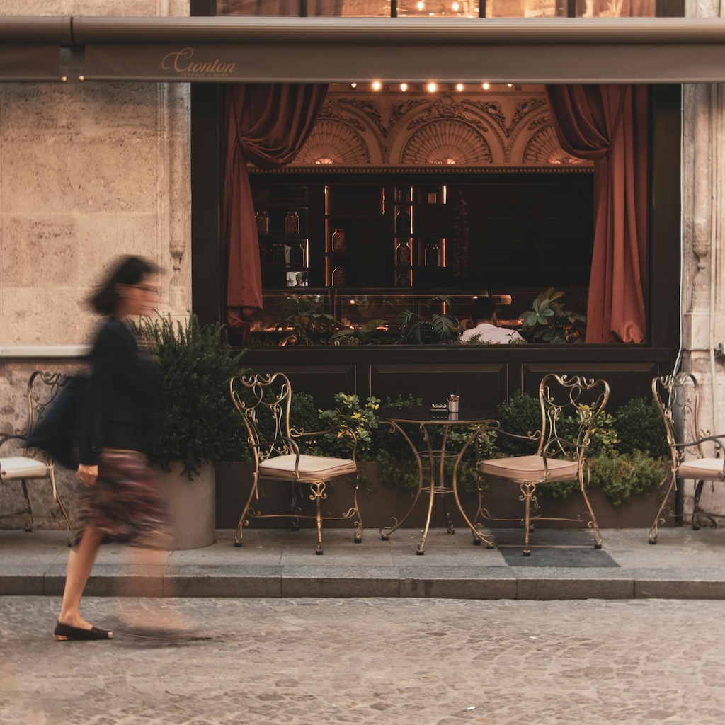 Charming outdoor seating area of a vintage café, featuring ornate wrought iron chairs and a small round table adorned with greenery, with a blurred motion of a person walking by in the foreground.