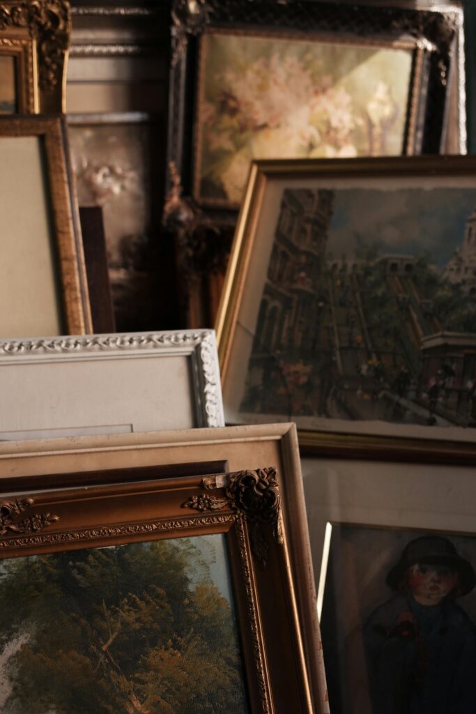 A collection of vintage framed artworks stacked together, showcasing intricate gold and silver frames with delicate details, highlighting a mix of floral and cityscape paintings.