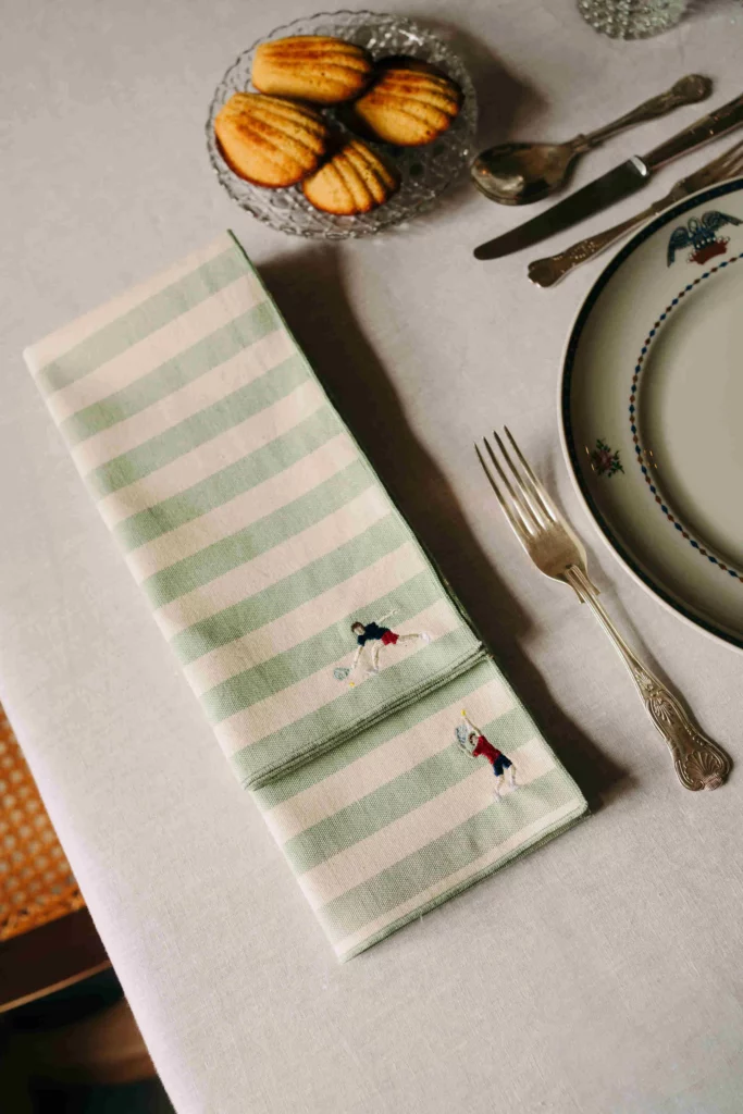 Embroidered Barra Napkin Green by Vintelio, green and white striped cotton napkin with embroidered tennis players, placed on a formal dining table with vintage plate and silverware, perfect for elegant entertaining