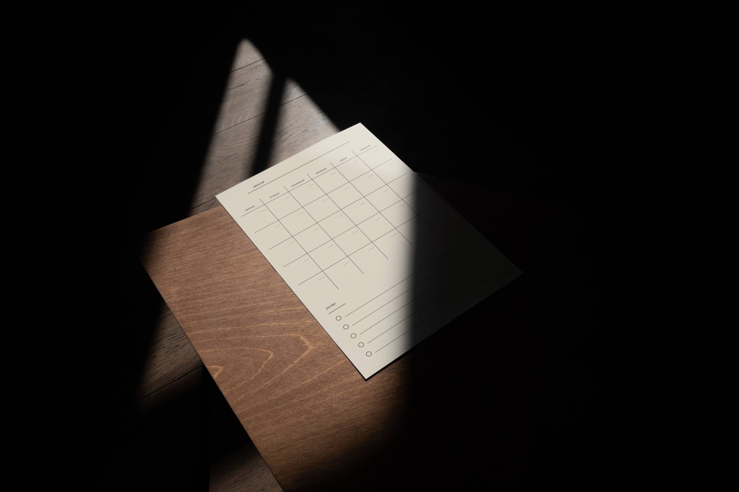 Minimalist beige magnetic planner with a monthly grid and “To Do” list, placed on a wooden surface in dramatic, partial sunlight.