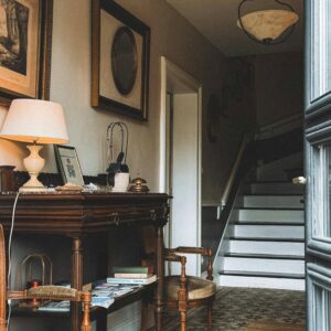 Vintage-inspired hallway featuring a dark wood console table with a white table lamp, framed art and mirrors on the wall, two upholstered antique chairs, patterned tile flooring, a woven rug, and a staircase in the background, creating a warm and elegant entryway.