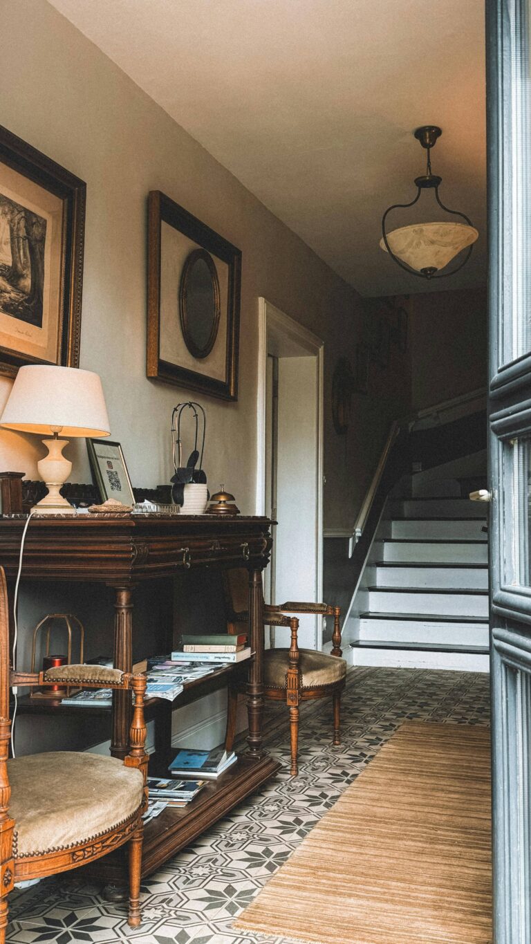 Vintage-inspired hallway featuring a dark wood console table with a white table lamp, framed art and mirrors on the wall, two upholstered antique chairs, patterned tile flooring, a woven rug, and a staircase in the background, creating a warm and elegant entryway.