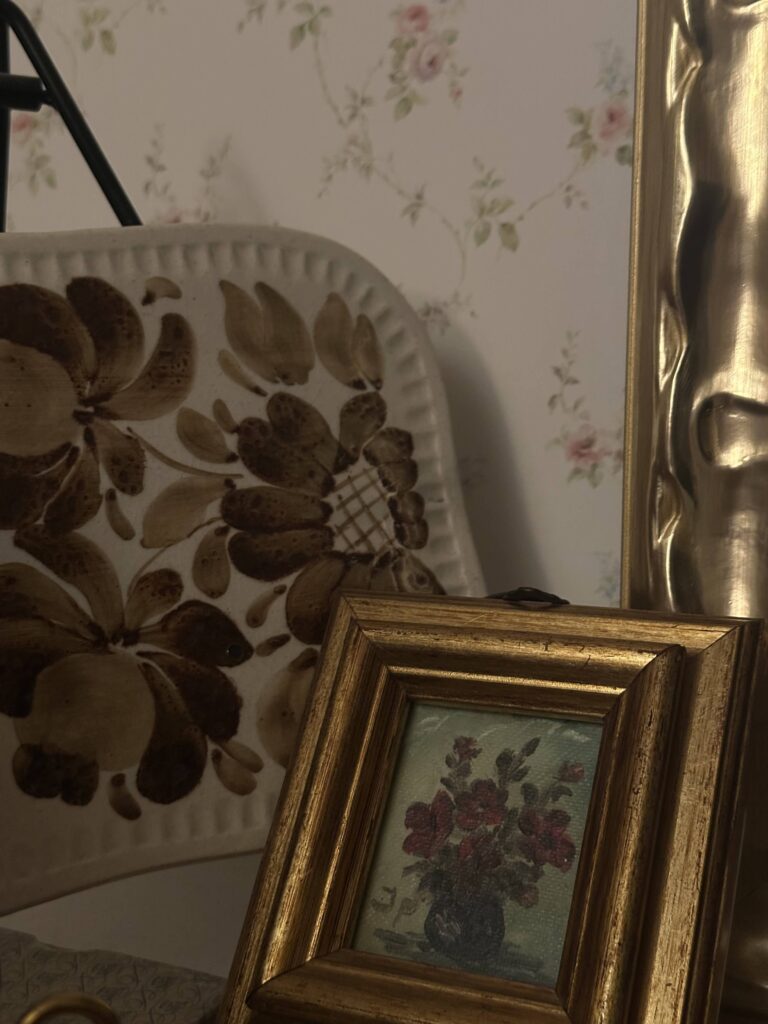 Beautiful composition of vintage accessories for home styling, featuring a gold ornate picture frame with a floral painting, a decorative ceramic plate with brown floral motifs, and a partial view of an elegant metal sculpture, all set against a soft floral wallpaper. These classic vintage-style décor elements create a warm, timeless, and nostalgic ambiance, perfect for vintage-inspired interiors. Ideal for adding character and charm to living rooms, bedrooms, or entryways with a touch of vintage elegance.